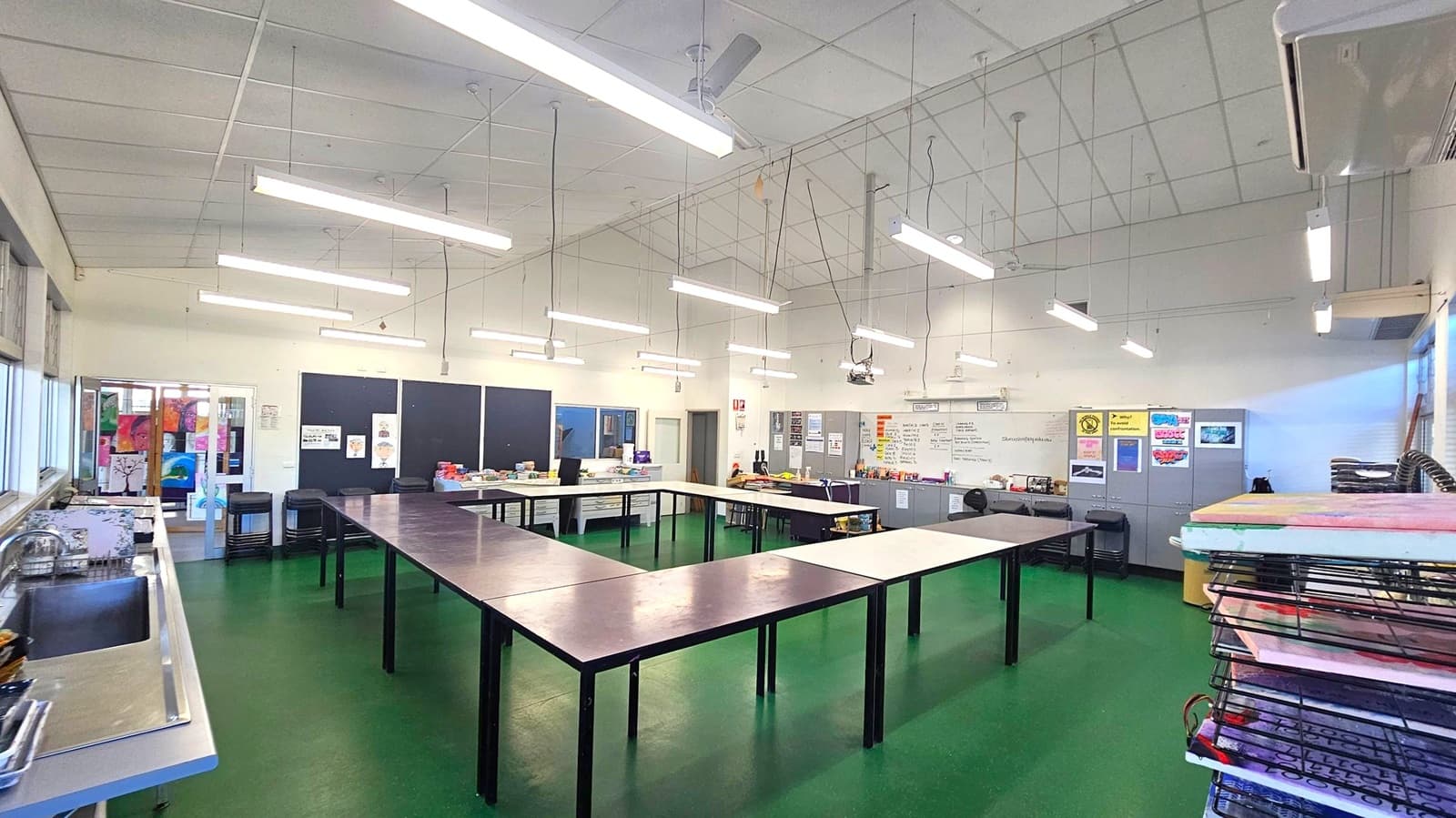 A classroom with a green floor and a white ceiling featuring fluorescent lights. In the centre, tables are arranged in a U-shape. The left side has a sink and windows, while the right side has stacked drying racks. The back wall displays posters and a whiteboard, with cabinets and various supplies. A projector hangs from the ceiling.