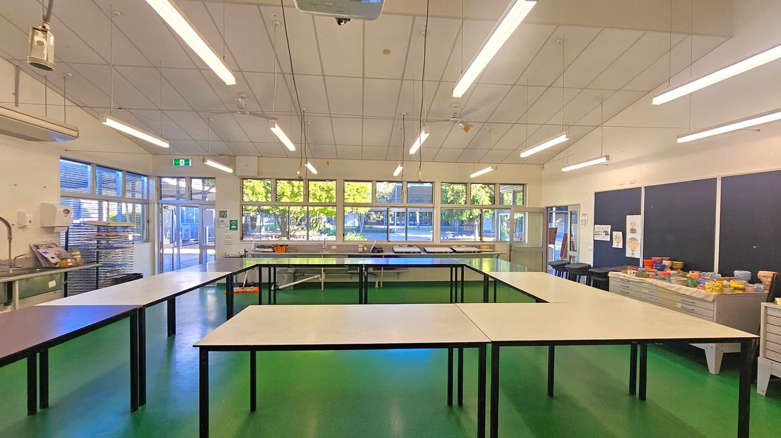 A spacious room with a green floor and a white ceiling featuring fluorescent lights. In the centre, there are several rectangular tables arranged in a U-shape. The left side has a sink and shelves with stacked trays. The right side has a long counter with various items and blackboards above. The back wall is lined with large windows and a glass door, offering a view of trees outside.