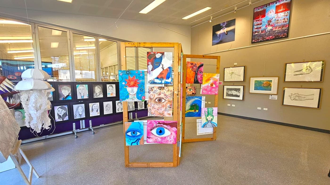 Art gallery with various artworks displayed. In the centre, a wooden stand holds colourful paintings, mainly featuring eyes and abstract faces. On the left, a series of black and white portraits are mounted on a black panel, with a white jellyfish sculpture nearby. The right wall displays framed drawings and paintings, including a swimmer and abstract designs. The floor is grey, and the ceiling has fluorescent lights.