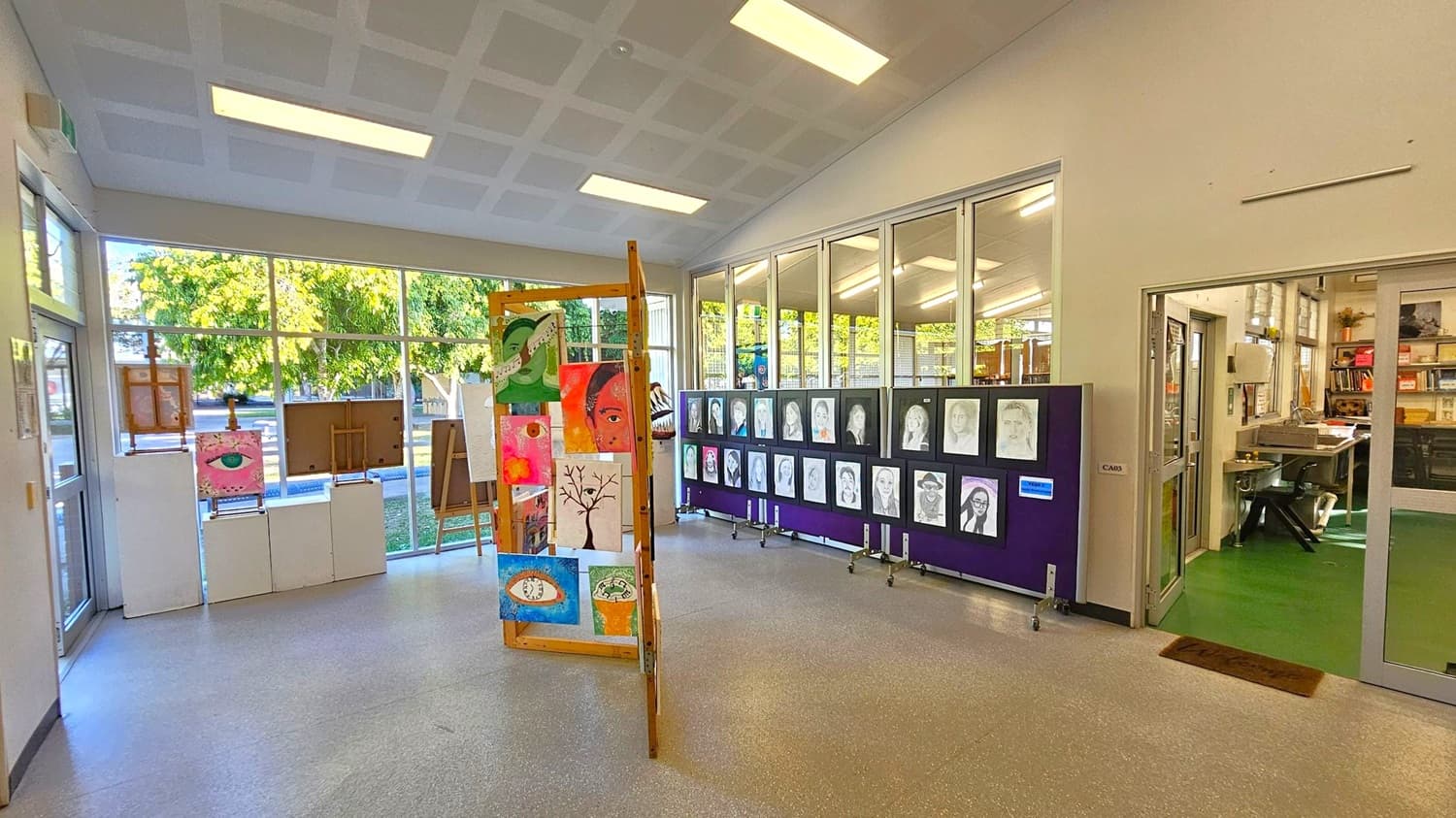 An art display area with various artworks. On the left, there are paintings on easels and stands near large windows showing trees outside. In the centre, a wooden stand holds multiple colourful paintings. On the right, a series of black and white portraits are mounted on a board. Beyond this, a room with green flooring and shelves is visible through glass doors. The ceiling is white with rectangular lights, and the floor is grey.