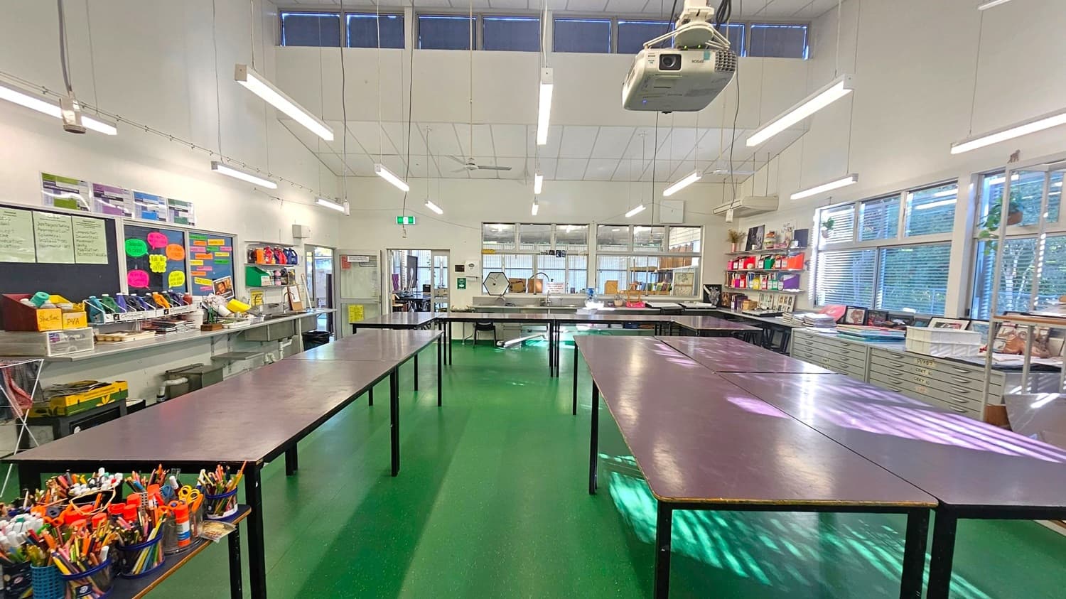 A spacious classroom with a green floor and high ceiling. In the foreground, several tables are arranged with art supplies like pencils and scissors. The left wall features shelves with colourful materials and a bulletin board with papers. The right side has large windows with blinds, allowing natural light in. The back wall has more tables and a door. A projector is mounted on the ceiling, and fluorescent lights are evenly spaced across the room.
