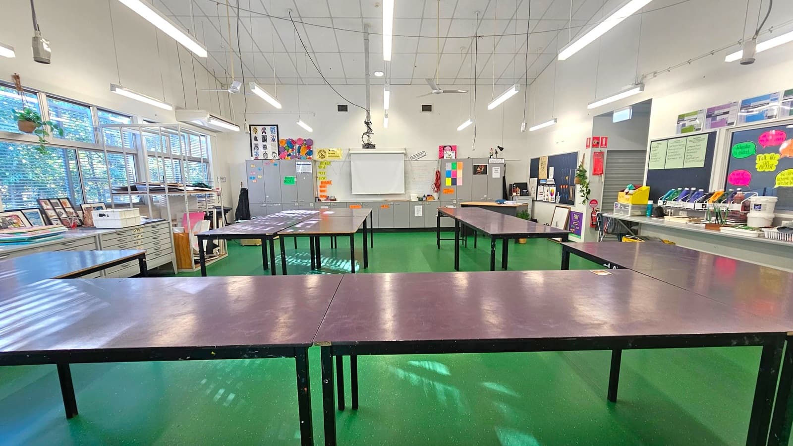 A classroom with green flooring and a white ceiling featuring fluorescent lights and hanging wires. In the centre, there are several large tables arranged in a U-shape. The left side has windows with blinds, shelves, and art supplies. The back wall has a whiteboard, cabinets, and colourful posters. The right side has more shelves with art materials and posters on the wall.