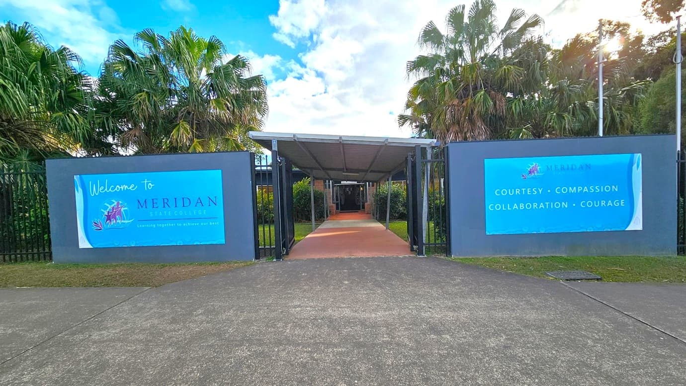 A pathway leads to a covered entrance flanked by two large signs. The left sign reads "Welcome to Meridan State College" with a logo and slogan. The right sign lists values: "Courtesy, Compassion, Collaboration, Courage." Palm trees are visible behind the signs. The ground is paved with concrete, and the entrance is framed by black metal fencing.