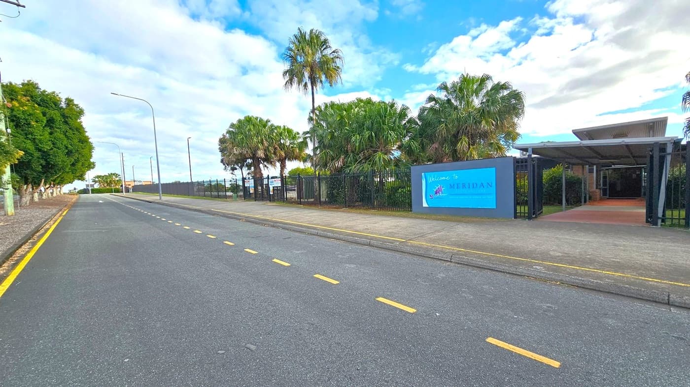 A road with a yellow line runs along the left side, bordered by a row of trees. On the right, a sign reads "Welcome to Meridan" near a gated entrance with palm trees behind it. The pavement is grey, and the sky is partly cloudy. Streetlights line the road, and a building with a slanted roof is visible in the background.