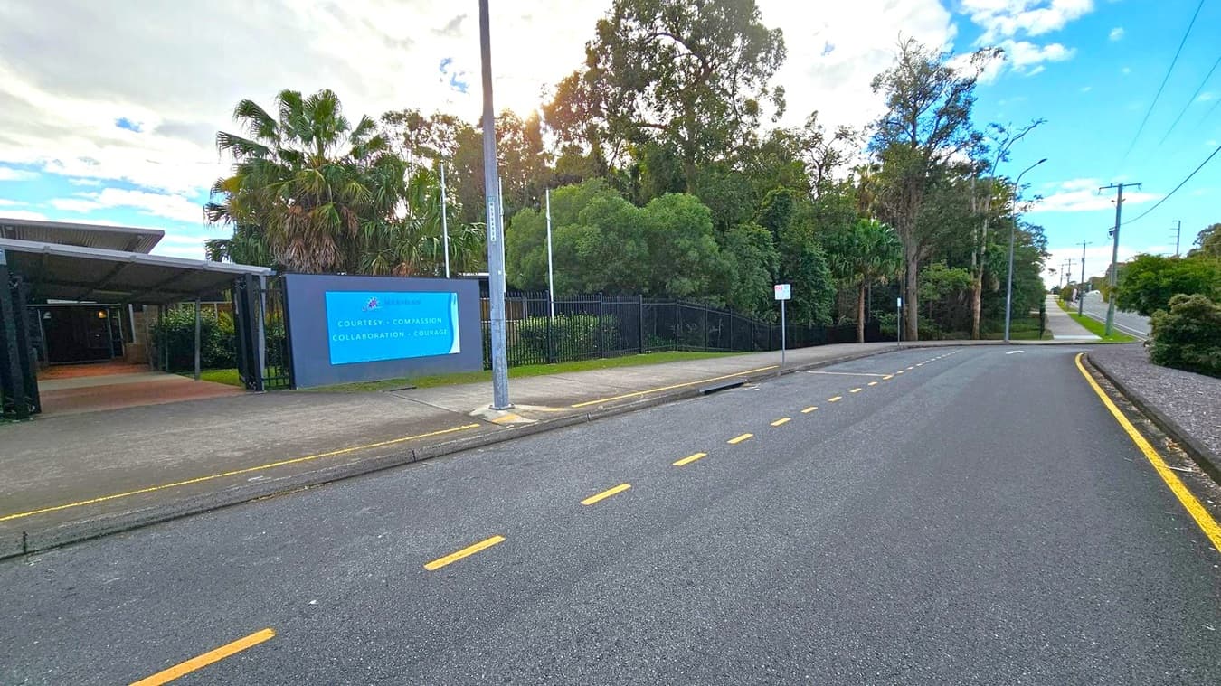A road curves gently to the right, bordered by a pavement with yellow markings. On the left, a building entrance is partially visible, with a blue sign displaying text. Tall palm trees and dense greenery are behind a black metal fence. A streetlamp stands near the sign. The sky is partly cloudy, and the sun is peeking through the trees, casting shadows on the pavement. The road continues into the distance, lined with more trees and utility poles.
