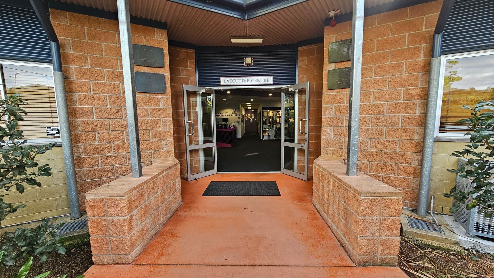 Entrance to a building with a sign reading "Executive Centre" above double glass doors. The walls are made of light brown brick, and there are metal poles on either side of the entrance. A black doormat lies on the reddish concrete floor leading inside. On the left, a window with blinds and a small plant; on the right, another window and a bush. Inside, a hallway with visible signage and displays. The ceiling is corrugated metal.