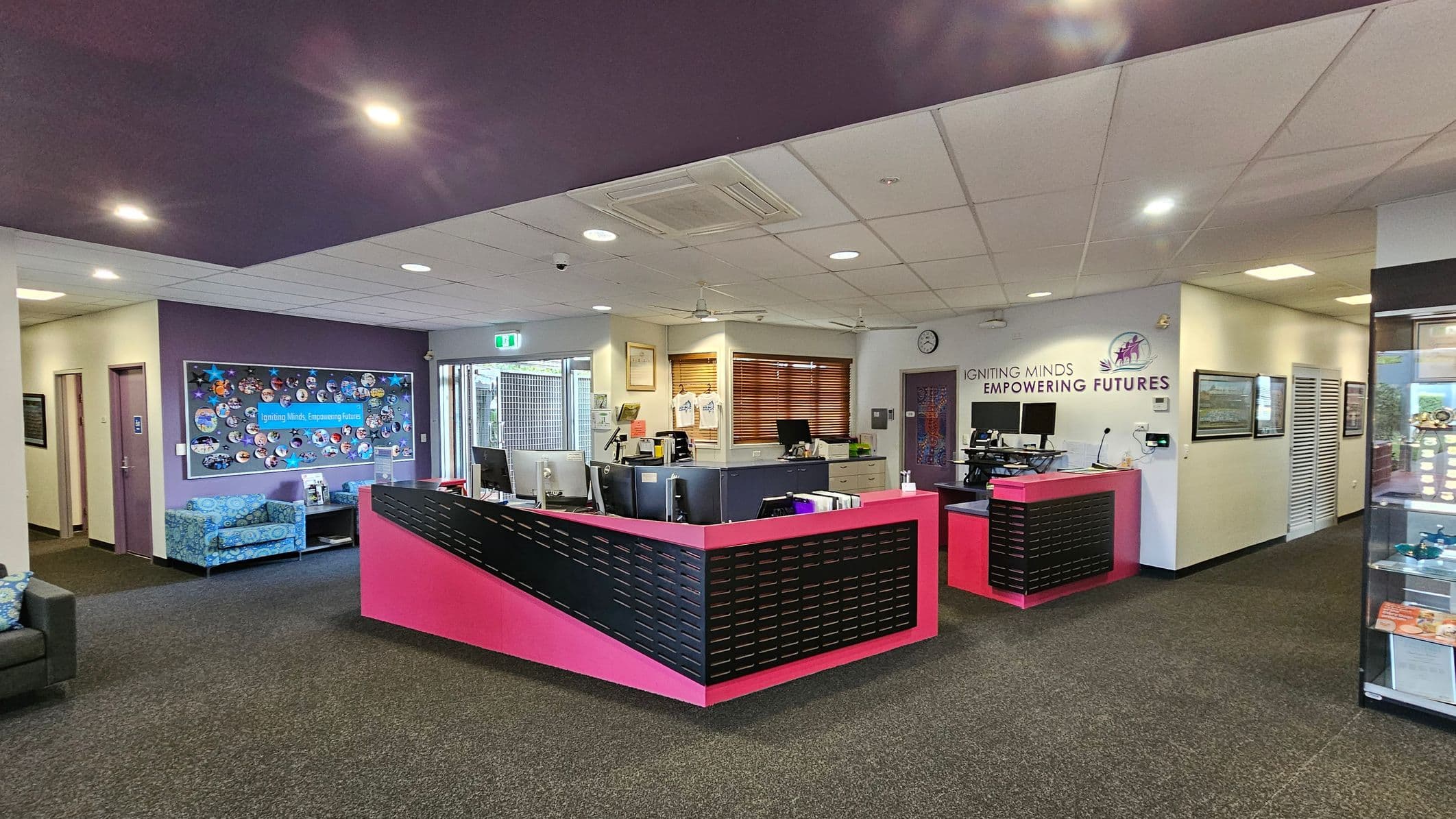 A reception area with a prominent pink and black desk in the centre, equipped with computers and office supplies. To the left, a seating area with blue patterned chairs and a colourful bulletin board on a purple wall. The right side features a display cabinet with various items. The back wall has a sign reading "IGNITING MINDS EMPOWERING FUTURES" and a clock. The ceiling is a mix of purple and white panels with recessed lighting, and the floor is carpeted in dark grey.