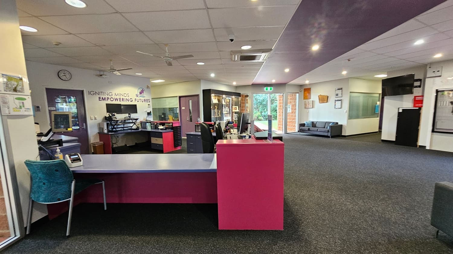 A spacious office area with a pink reception desk in the foreground. To the left, a teal chair is positioned near the desk. The back wall features the words "IGNITING MINDS EMPOWERING FUTURES" and a clock. Several desks with computers and office supplies are visible. The right side has a grey sofa against the wall, and a glass door leading outside. The ceiling is fitted with lights and air conditioning vents, and the floor is carpeted in a dark colour.