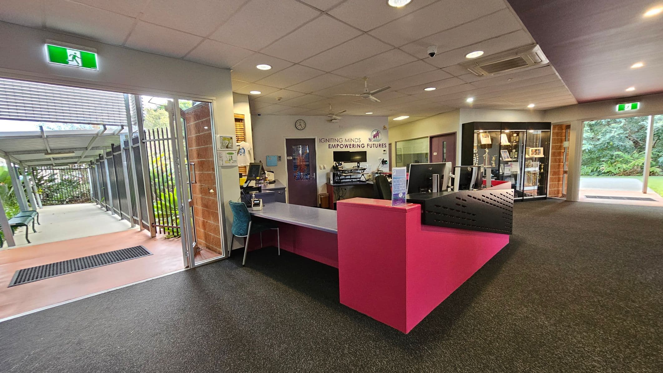 A reception area with a pink desk in the centre, equipped with computers and a chair. To the left, a glass door opens to an outdoor walkway with benches and greenery. On the right, a display cabinet with various items is visible. The wall behind the desk features the words "IGNITING MINDS EMPOWERING FUTURES." The ceiling has recessed lighting and a fan, while the floor is carpeted in dark grey. An exit sign is above the doorways.