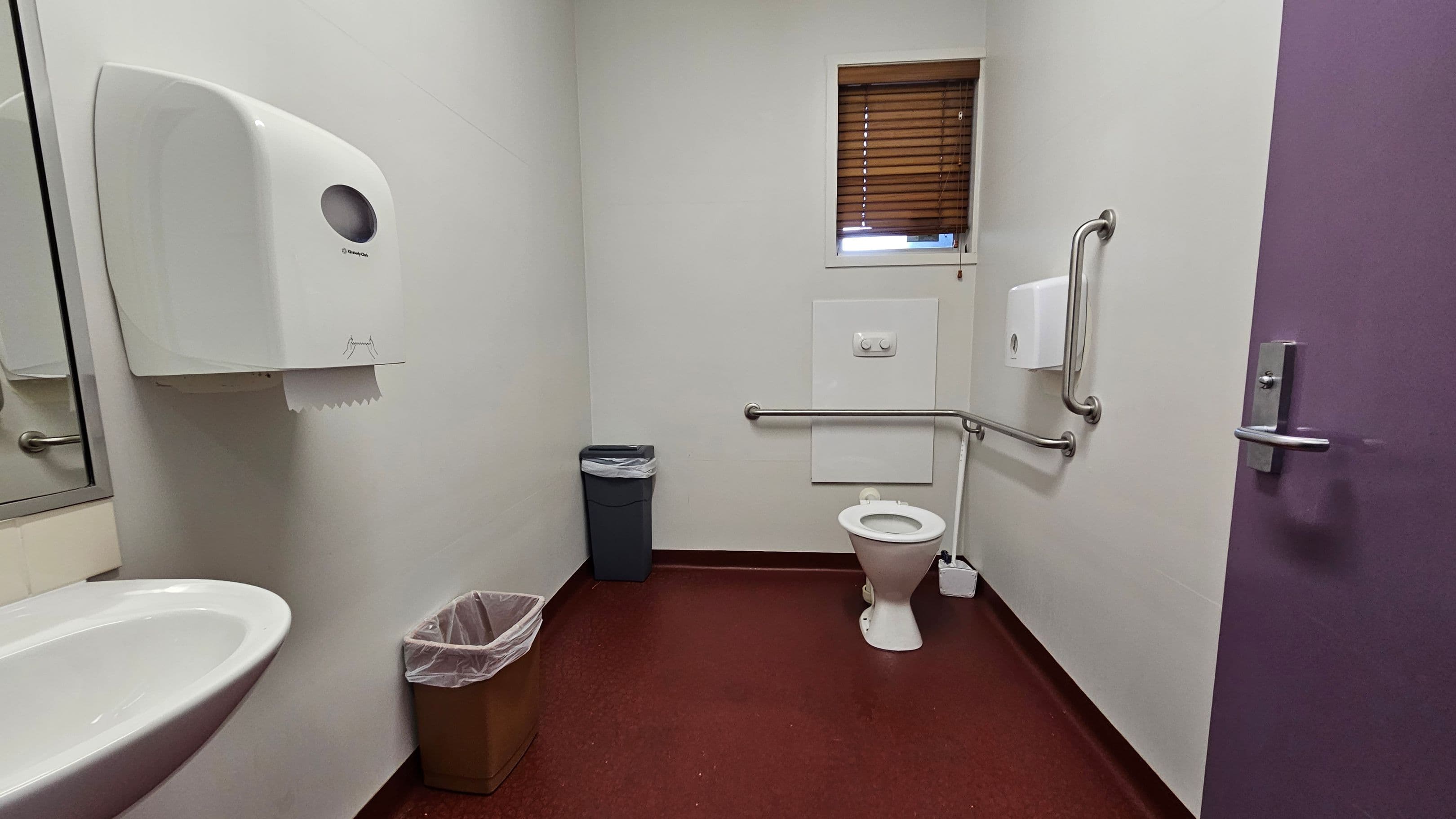 A bathroom with a red floor and white walls. On the left, a white sink with a mirror above it and a paper towel dispenser. In the centre, a grey bin and a brown bin with a liner. On the right, a white toilet with a grab rail beside it and a small window with wooden blinds above. A purple door with a metal handle is partially visible.
