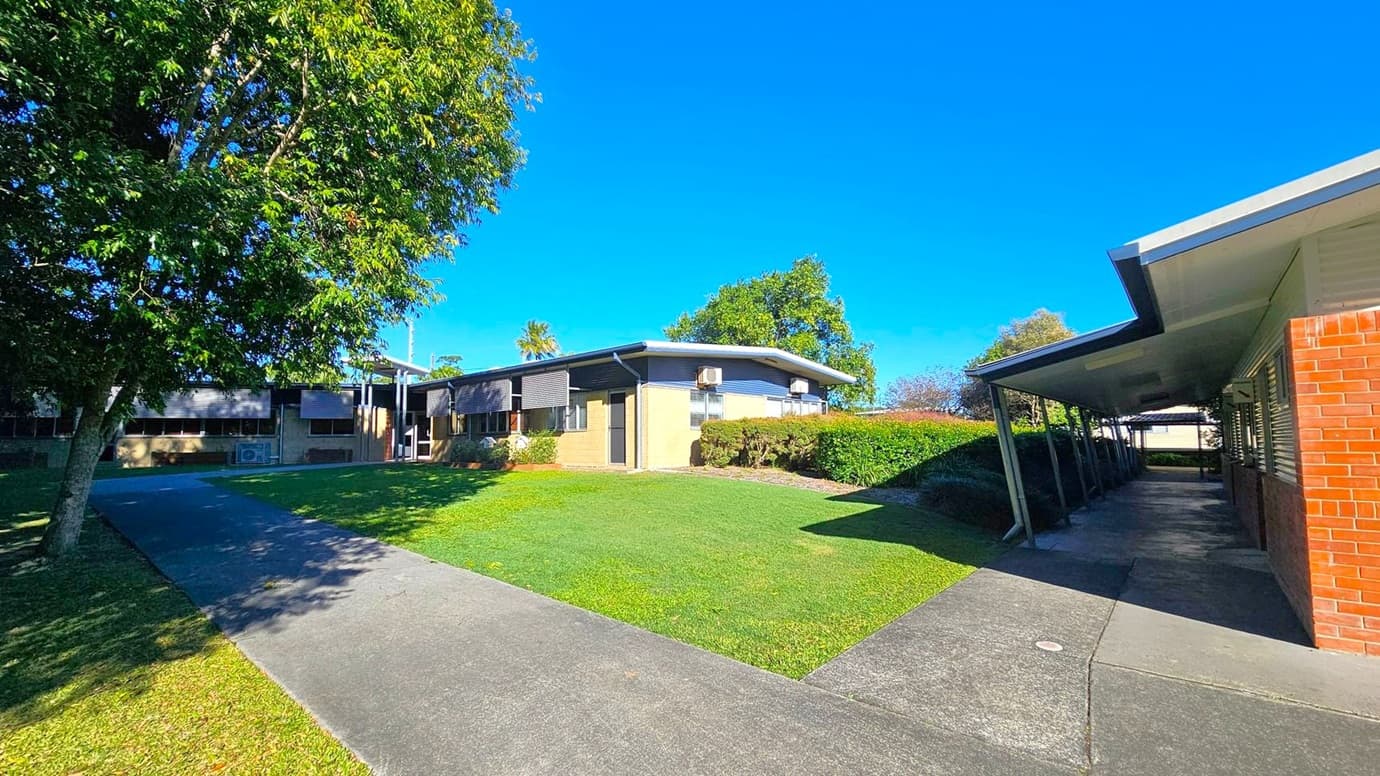 A grassy courtyard is surrounded by single-storey buildings. On the left, a large tree provides shade over a concrete path. In the centre, a building with a sloped roof and air conditioning units is visible, with hedges lining its base. On the right, a covered walkway with a red brick wall leads into the distance. The sky is clear and blue.