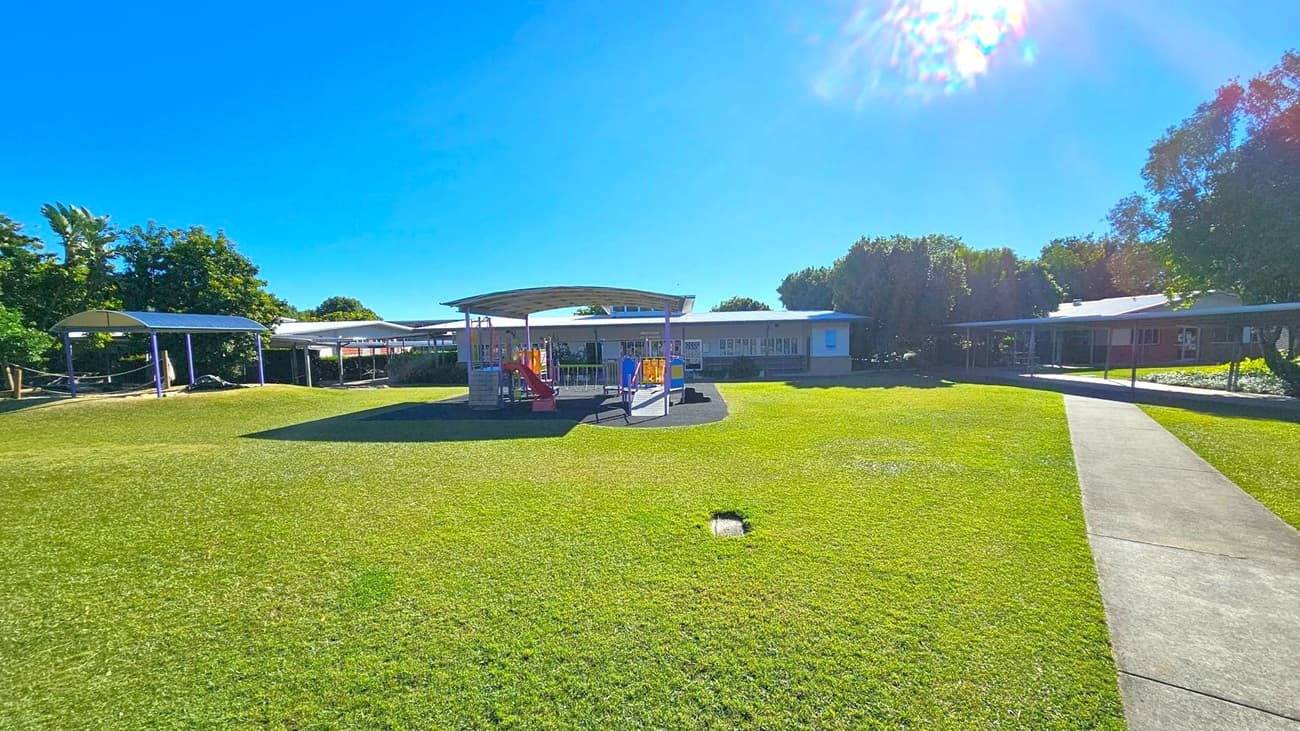 A grassy field with a playground structure in the centre, featuring slides and climbing equipment under a metal roof. To the left, there is a covered walkway and trees. On the right, a concrete path leads towards a building with a long, shaded veranda. Trees are visible in the background, providing shade and greenery. The sky is clear and blue.