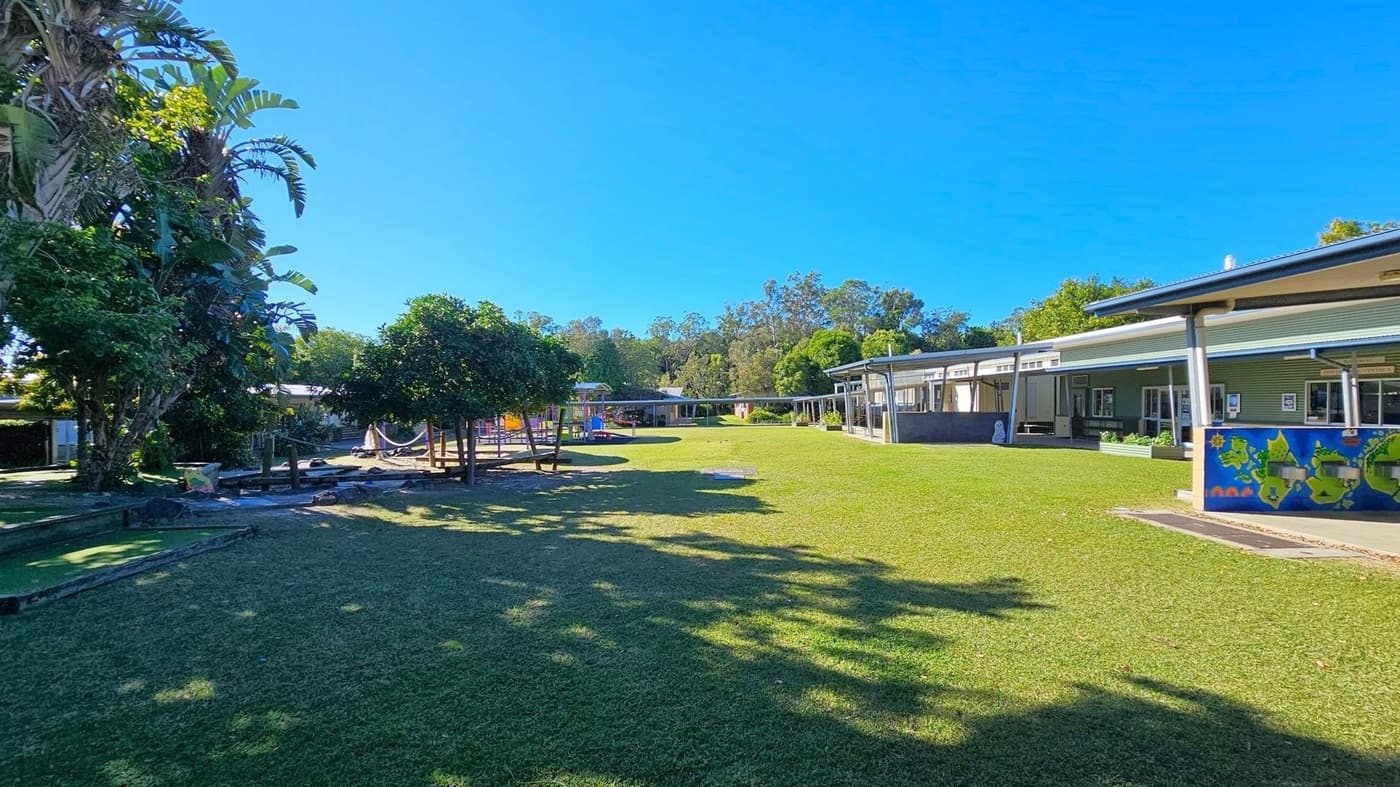 A grassy open area with a playground in the centre, featuring colourful equipment. On the left, there are tall trees with dense foliage. To the right, a building with a covered walkway and a mural on the wall. The background has more trees and additional structures. Shadows from the trees are cast on the grass.