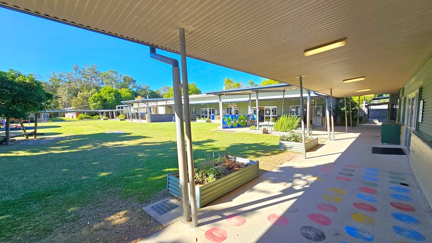 A covered walkway with a corrugated metal ceiling and fluorescent lights runs along the right side, with a building featuring large windows. The floor has colourful circular patterns. On the left, there's a grassy area with trees and a playground. In the background, more buildings with metal roofs and a mural are visible, surrounded by greenery. Raised garden beds are near the walkway.