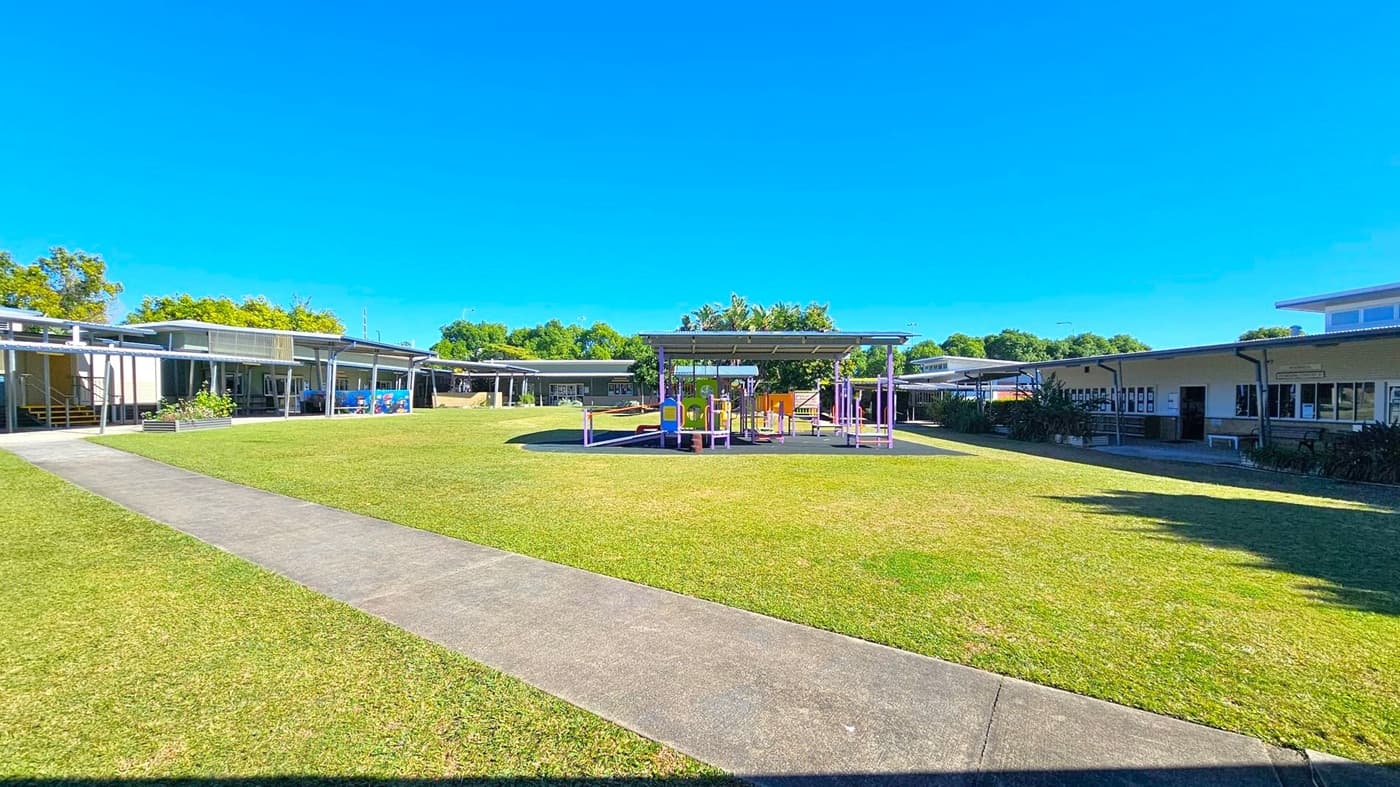 A grassy courtyard with a paved path running through it. In the centre, there's a playground structure with purple and yellow elements, covered by a roof. Surrounding the courtyard are single-storey buildings with large windows and metal roofs. Trees and shrubs are visible in the background, providing greenery around the area.