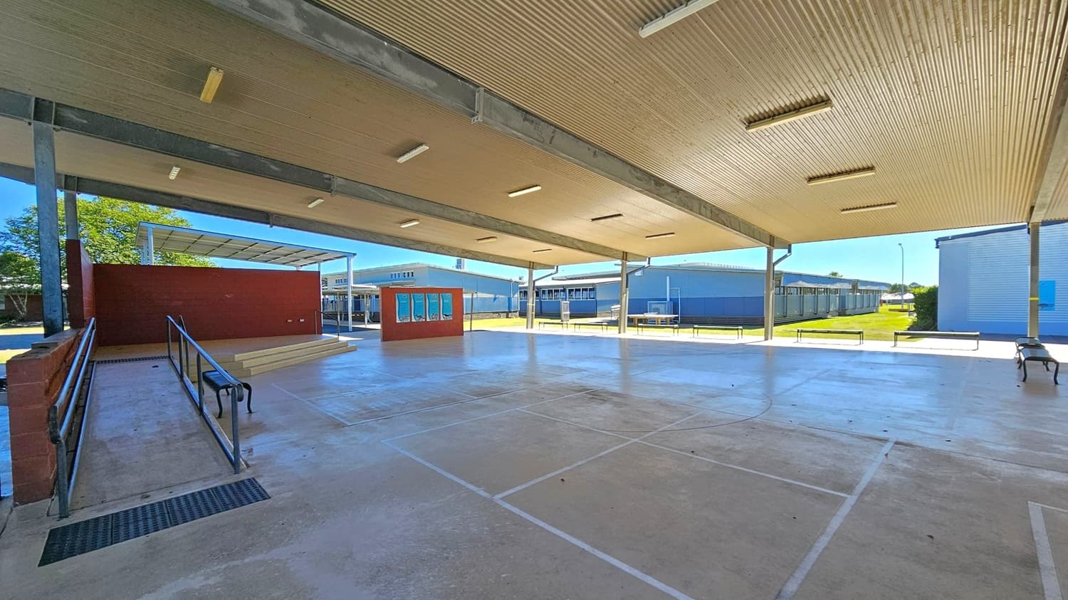 A covered outdoor area with a corrugated metal ceiling and fluorescent lights. The floor is concrete with faint markings. On the left, there is a ramp with metal railings leading to a small stage with a red brick backdrop. In the centre, a red wall with posters is visible. The background shows several single-storey buildings with metal roofs, and a grassy area. A bench is on the right side.