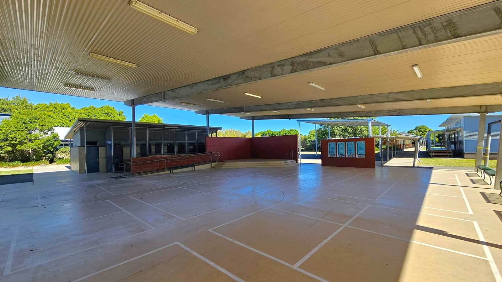 A covered outdoor area with a corrugated metal ceiling and fluorescent lights. The floor is marked with sports lines. On the left, there is a building with a blue door and windows, and a row of red benches. In the centre, a maroon wall with steps leads to an open space. On the right, a noticeboard is visible, and beyond it, a pathway leads to another building. Trees and greenery are visible in the background.
