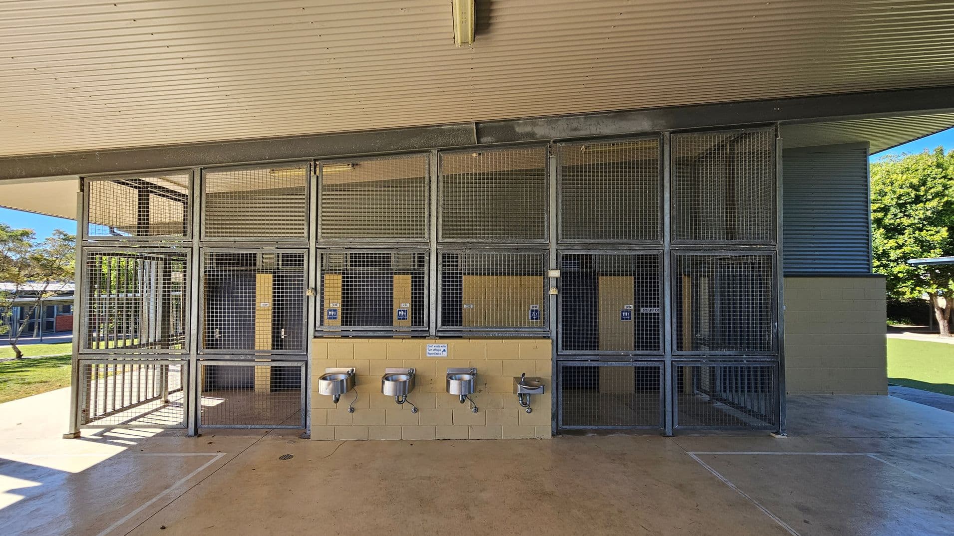 A structure with a corrugated metal roof and wire mesh walls. In the centre, there are four stainless steel drinking fountains mounted on a beige brick wall. Behind the mesh, there are doors with signs. The floor is concrete, and there is a grassy area with trees visible in the background on the left and right sides.