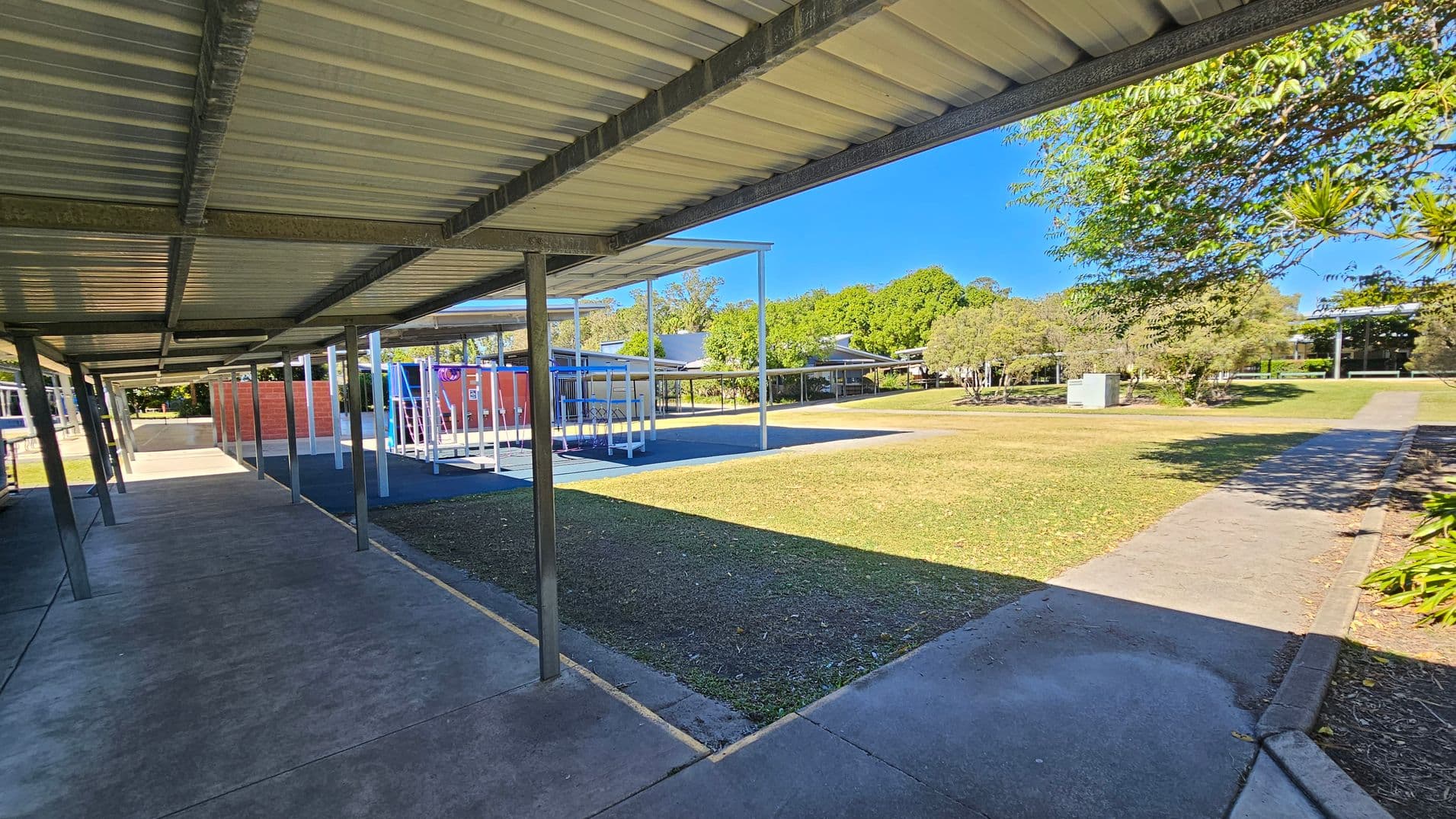 A covered walkway with a metal roof and supporting beams runs along the left side. To the right, there is a grassy area with a paved path leading through it. In the middle, a playground with blue and red equipment is visible. Trees and shrubs are scattered throughout the background, with buildings partially visible beyond them. The ground is a mix of concrete and grass, and the area is well-lit by sunlight.
