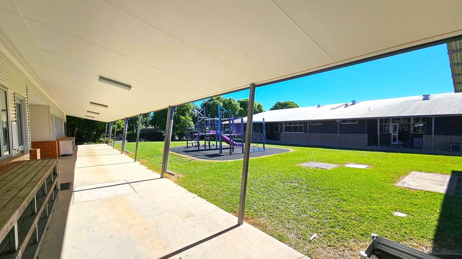 A covered walkway with a white ceiling and fluorescent lights runs along the left side, adjacent to a building with windows and a brick section. On the right, a grassy area features a playground with purple and grey equipment. In the background, another building with a corrugated metal roof is visible. The floor is concrete, and metal poles support the roof of the walkway.