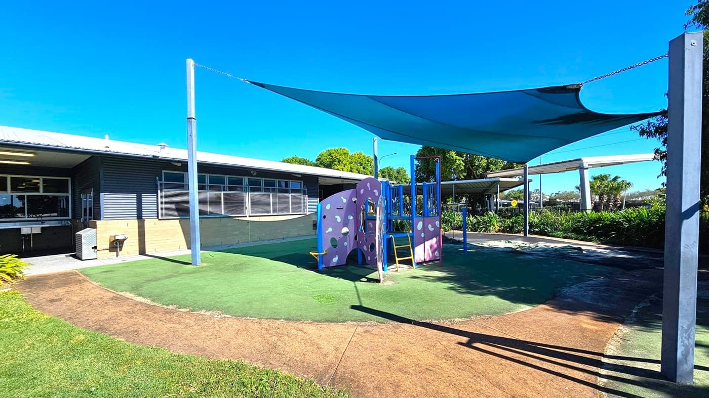 A playground with a purple climbing structure and yellow ladder is in the centre, set on artificial grass. A large blue shade sail is overhead, supported by metal poles. To the left, a building with a corrugated metal roof and windows is visible. The background features lush greenery and a covered walkway. The ground is a mix of green artificial turf and brown pathways.