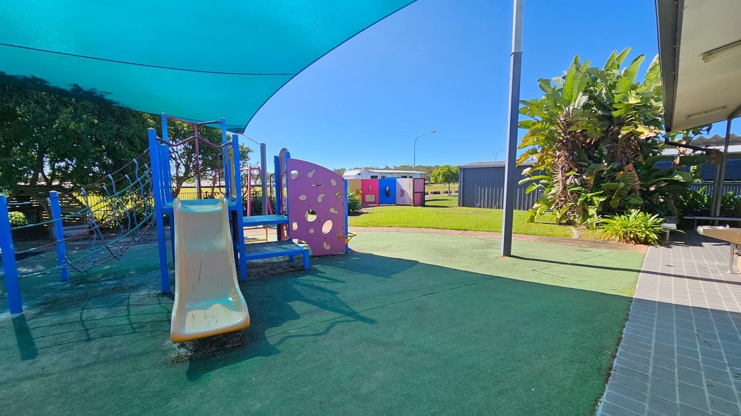 A playground with a blue shade sail overhead. On the left, there's a play structure with a yellow slide, blue steps, and a climbing net. In the middle, a purple panel with circular cutouts. The ground is covered with green safety matting. To the right, a large plant with broad leaves and a paved area. In the background, a small building and a grassy area with a fence.