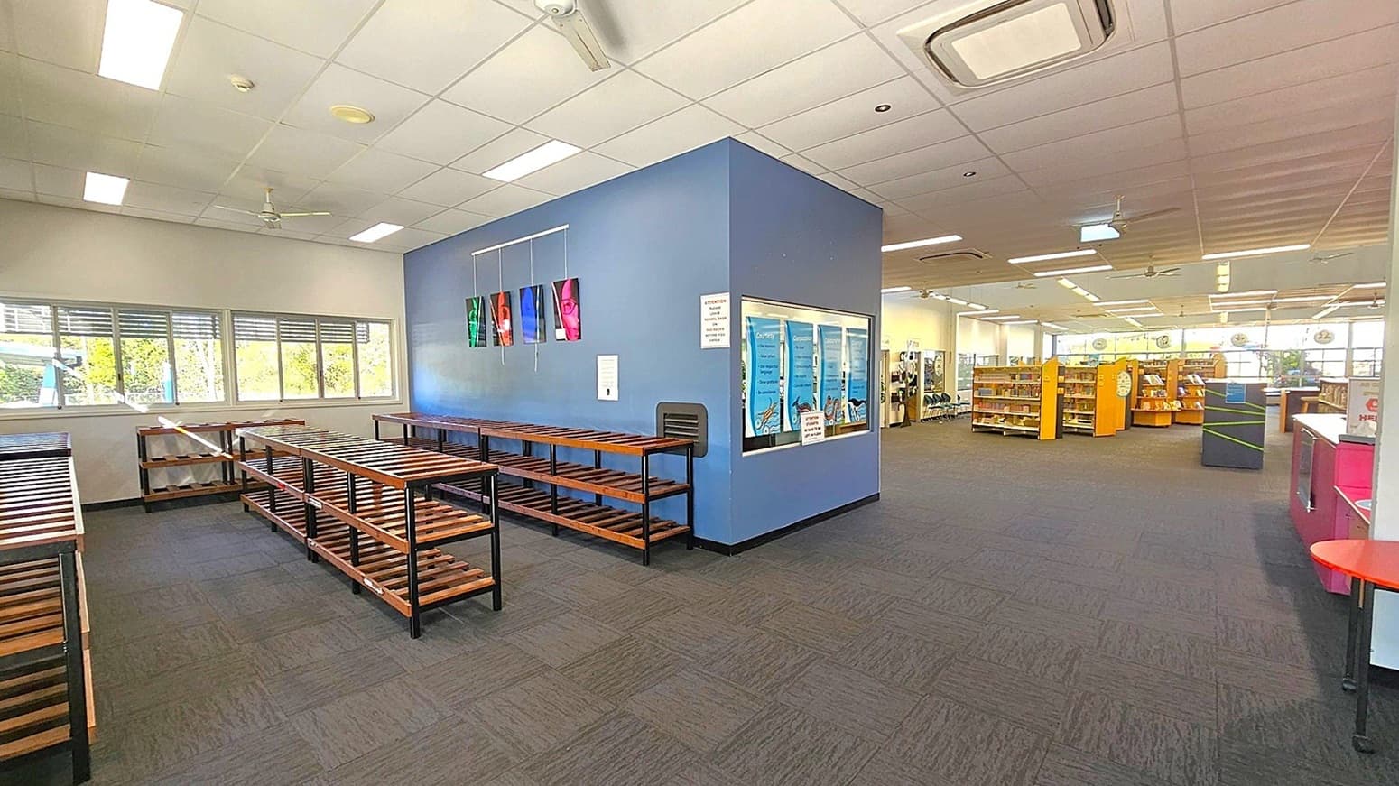 A library interior with a blue accent wall in the centre, displaying colourful vertical banners. To the left, there are empty wooden shelves and large windows with blinds. On the right, there are bookshelves filled with books and a help desk with a red counter. The floor is carpeted in dark grey with patches of green. The ceiling is white with fluorescent lights and air conditioning vents.