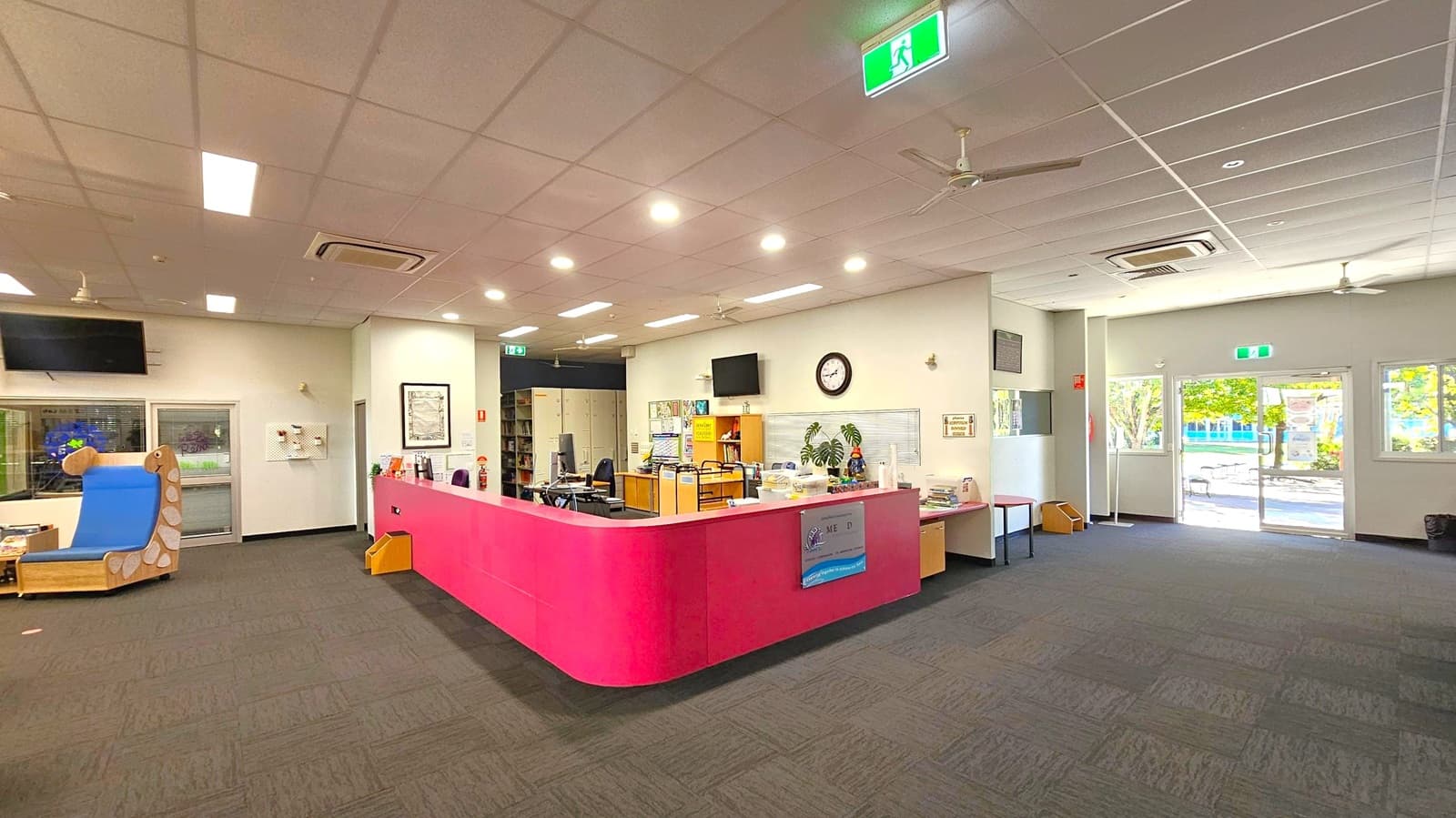 A spacious room with a dark carpeted floor and a white panelled ceiling featuring recessed lighting and ceiling fans. In the centre, a pink reception desk is equipped with office supplies and a clock on the wall behind it. To the left, a blue cushioned chair with a wooden frame is near a window. On the right, glass doors lead outside, allowing natural light to enter. An emergency exit sign is visible above the doors.