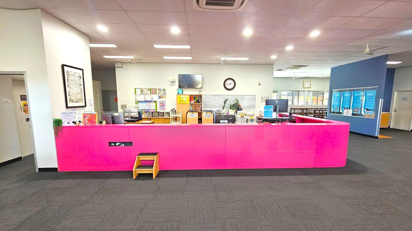 A reception area with a bright pink counter in the foreground, featuring a small wooden step stool. Behind the counter are various office items, including computers, a clock, and a television mounted on the wall. The background has a bulletin board with colourful posters and a plant. To the right, there is a blue wall with large posters and a door. The ceiling has recessed lighting, and the floor is covered with dark carpet tiles.