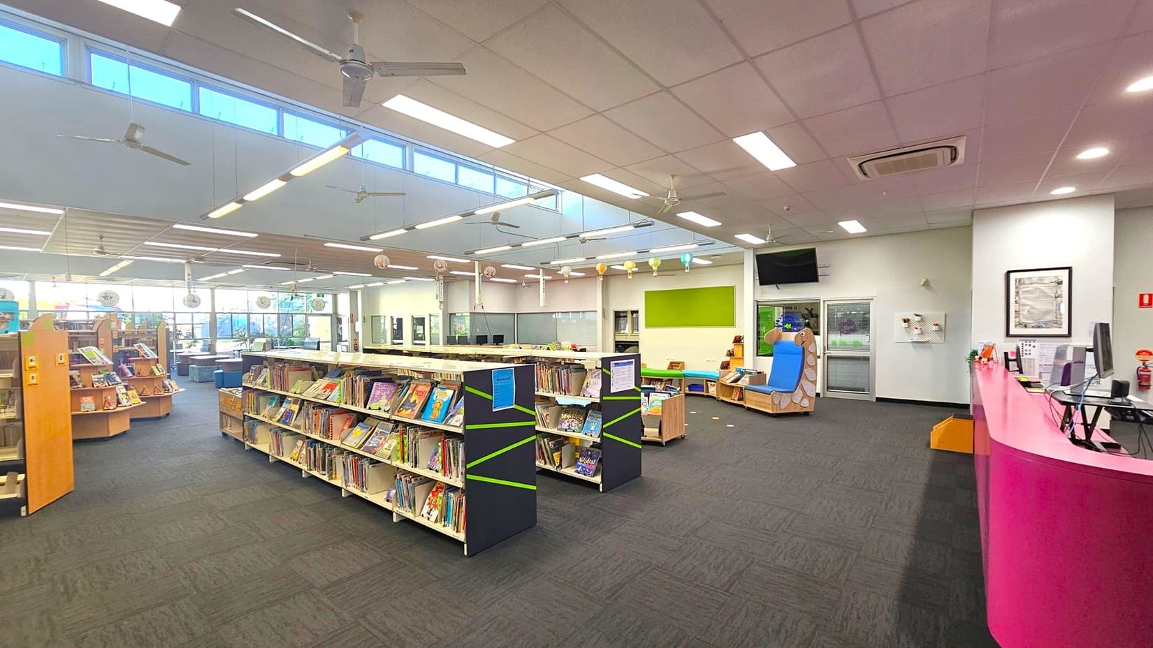 A spacious library interior with a high ceiling featuring fluorescent lights and ceiling fans. On the left, there are bookshelves filled with colourful books. In the centre, more bookshelves are arranged neatly. To the right, a pink reception desk with computers and a small seating area with a blue and wooden bench. The floor is carpeted in dark grey, and the walls are light-coloured with a green accent panel. Large windows in the background allow natural light to enter.