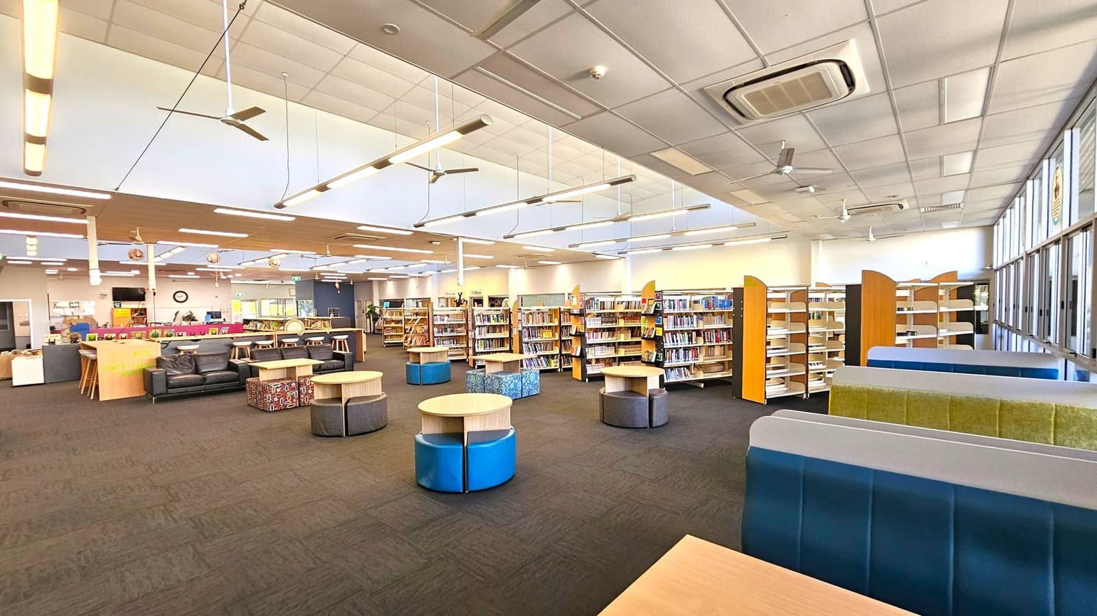 Spacious library interior with a dark carpeted floor. In the foreground, there are round tables with cushioned stools. To the right, there are blue and green upholstered seating booths. The middle area has several bookshelves filled with books. The left side features a seating area with black sofas and a counter with stools. The ceiling is fitted with fluorescent lights and ceiling fans. Large windows line the right wall, allowing natural light to enter.