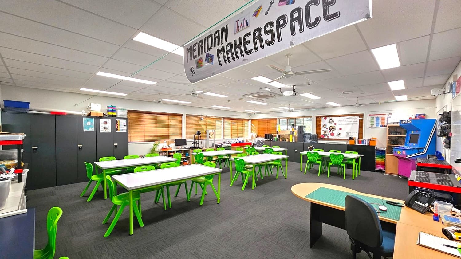 A classroom with a sign reading "Meridan Makerspace" hanging from the ceiling. The room features several tables with bright green chairs arranged around them. On the left, there are black storage cabinets and shelves with various supplies. The right side has a blue arcade machine labelled "Game On" and more storage units. The back wall has windows with wooden blinds and a variety of equipment and materials. The floor is carpeted in dark grey, and the ceiling has fluorescent lights.