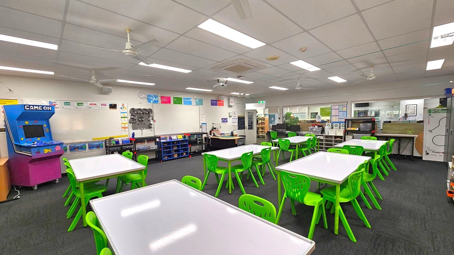 A classroom with several white tables and bright green chairs arranged in groups. On the left, there's a blue arcade machine labelled "Game On." The back wall features a whiteboard with various educational posters above it. Shelves with books and supplies are beneath the whiteboard. The right side has more storage units and a small kitchen area with a microwave. The ceiling is fitted with fluorescent lights and ceiling fans, and the floor is carpeted in dark grey.