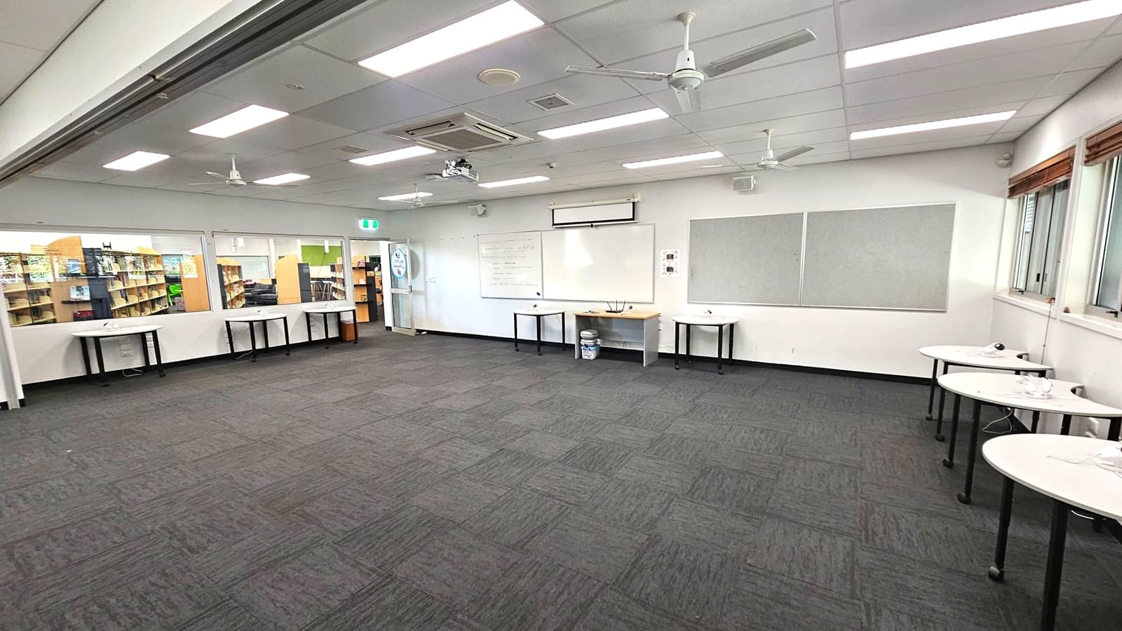 A spacious classroom with grey carpeted flooring and a white ceiling featuring fluorescent lights and ceiling fans. The back wall has a whiteboard and a bulletin board, with several tables placed against it. On the left, large windows reveal a library area with bookshelves. On the right, there are more tables with computers and windows with blinds. A door with a glass panel is visible near the centre-left, leading to the library.