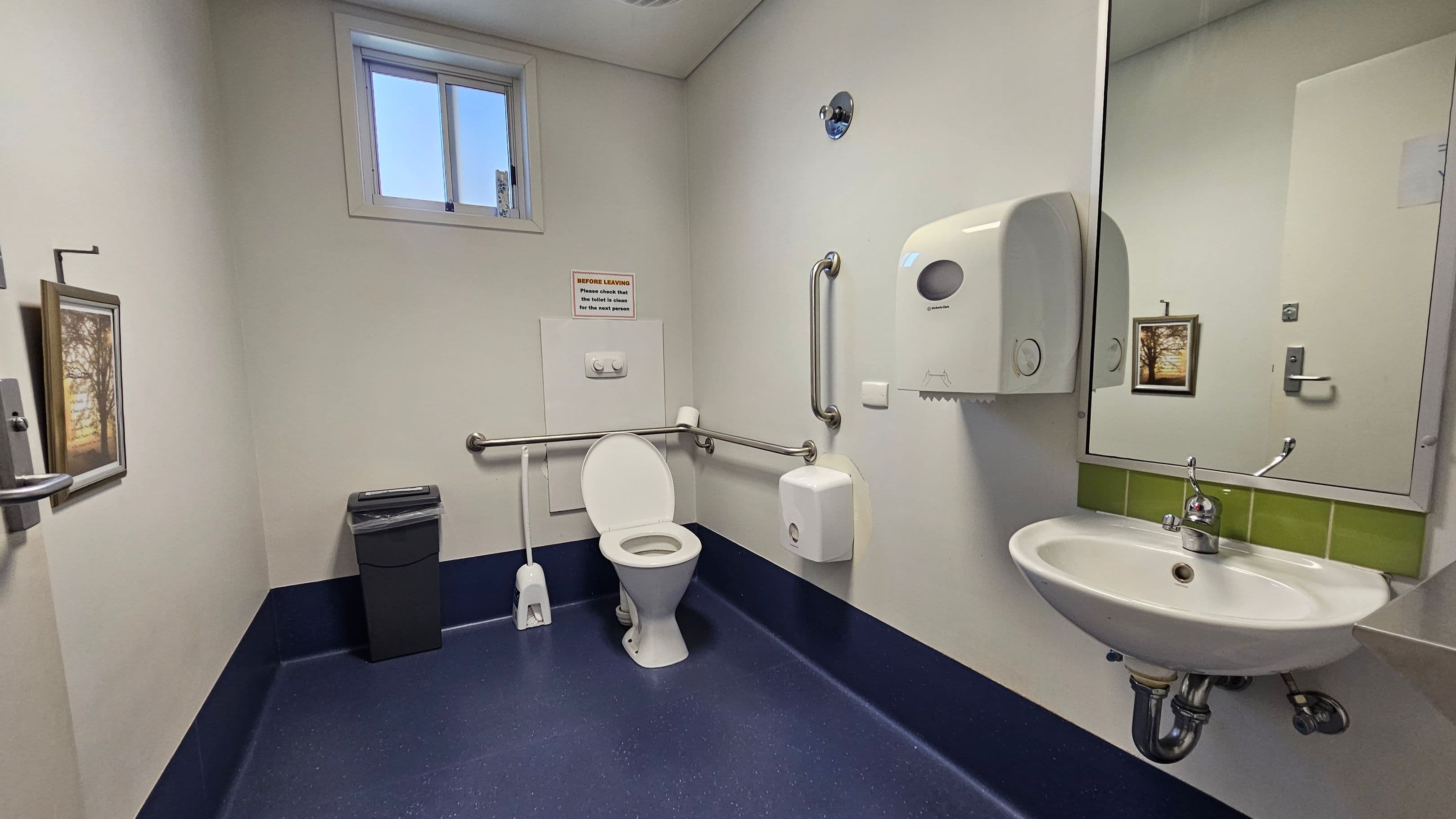 A bathroom with a white toilet in the centre, equipped with metal grab bars on both sides. To the right, a white sink with a mirror above it and a paper towel dispenser nearby. A small window is positioned high on the wall above the toilet. On the left, a waste bin and a framed picture are visible. The floor is dark blue, and the walls are light-coloured. A sign is mounted above the toilet.