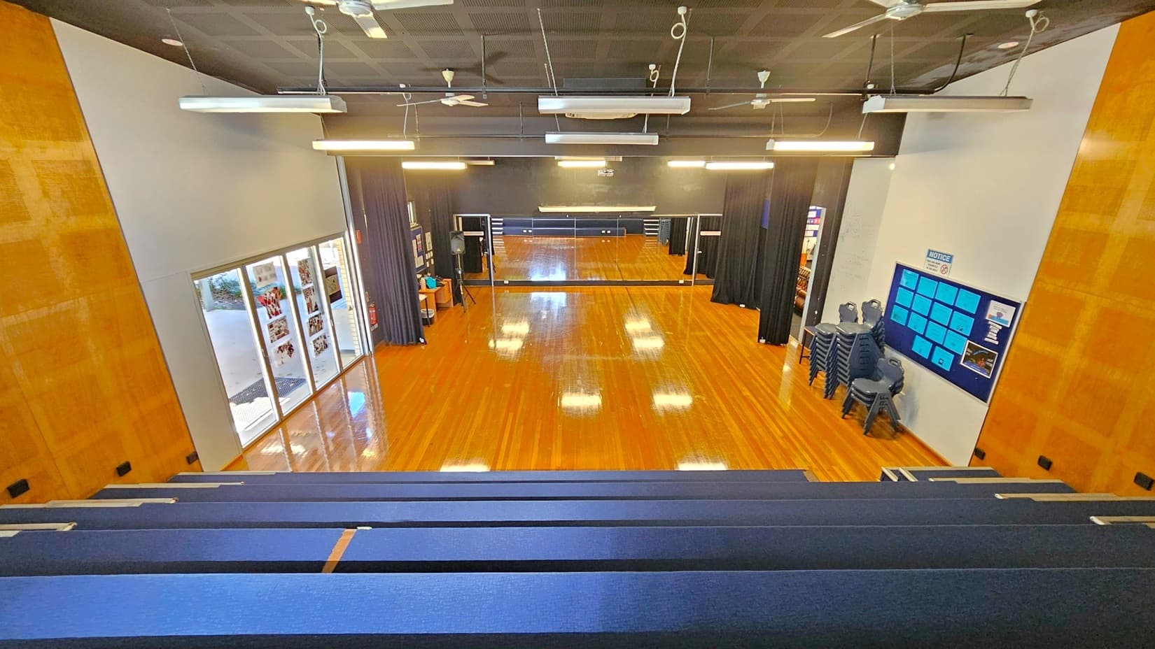 A spacious room with a polished wooden floor and a black ceiling featuring hanging lights and fans. On the left, there are large windows with posters displayed. The right side has a notice board with blue papers and a stack of chairs. In the centre, a large mirror spans the wall, reflecting the room. The foreground shows tiered seating with blue cushions. Black curtains frame the mirror, and the walls have a wooden panel design.