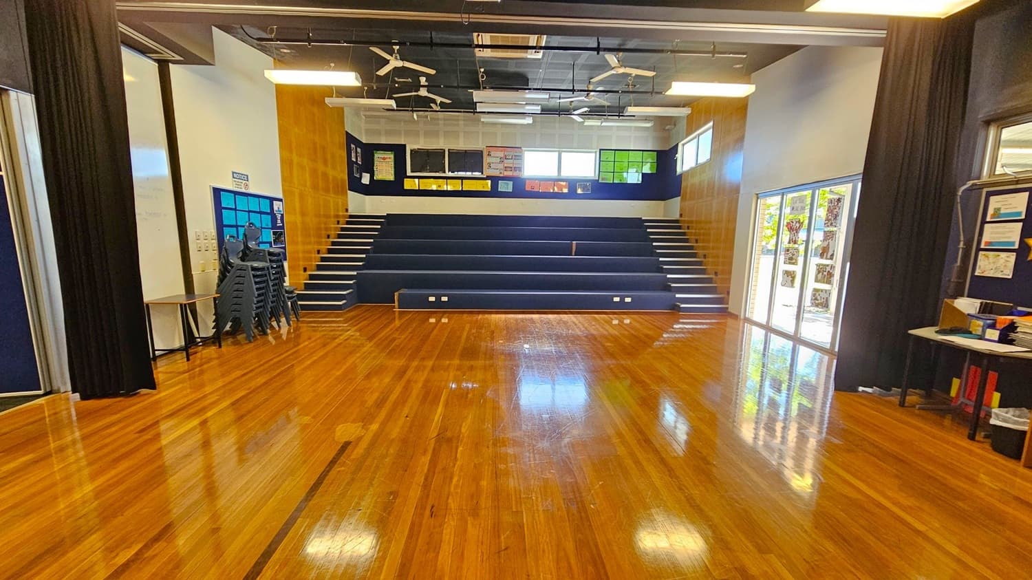 A spacious room with polished wooden floors and tiered seating at the back. On the left, there are stacked chairs and a notice board. On the right, large windows with glass doors let in natural light, and a table with various items is visible. The ceiling features exposed beams, lights, and ceiling fans. Walls are a mix of wood panelling and white surfaces, with colourful posters displayed at the back.