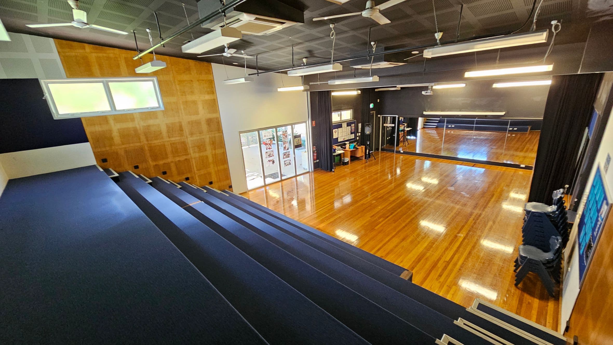 A spacious room with polished wooden floors and a high ceiling. On the left, there are tiered blue seating steps leading down to the floor. The right side features a stack of black chairs and a large mirror reflecting the room. The back wall has a wooden panel with a window, and there are glass doors leading to another area. The ceiling is equipped with lights, fans, and air conditioning units.