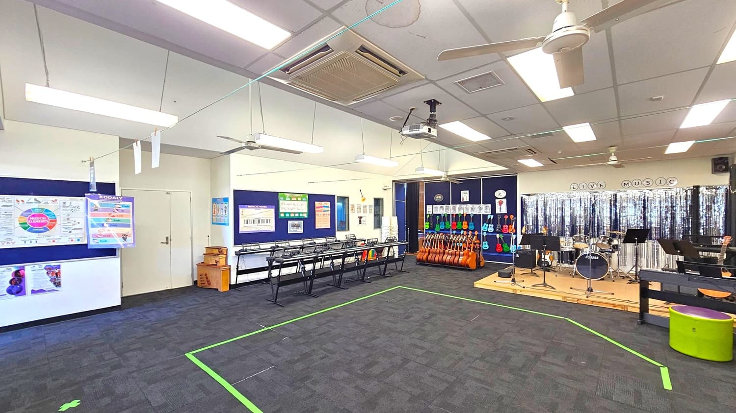A music classroom with a dark carpeted floor and a ceiling featuring fluorescent lights and ceiling fans. On the left, there are music theory posters and a row of black music stands. The centre has a collection of guitars and ukuleles against a wall. On the right, a small stage with a drum set and music stands is set up, with a backdrop of silver streamers. The words "LIVE MUSIC" are displayed above the stage area. A green line is marked on the floor.
