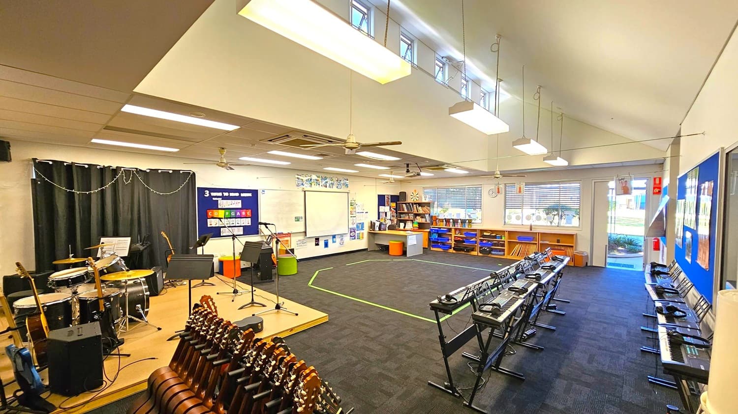 A music classroom with a stage on the left featuring a drum set, guitars, and music stands. The middle area has a large open space with a green tape outline on the carpet. On the right, there are rows of keyboards with headphones on stands. The back wall has a whiteboard, bookshelves, and a window with blinds. The ceiling is equipped with fluorescent lights and fans, and the floor is carpeted. A door on the right leads outside.