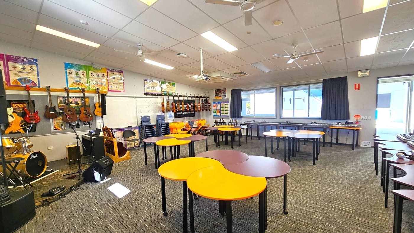 A classroom with a carpeted floor and a grid-patterned ceiling with fluorescent lights and ceiling fans. On the left, there are guitars hanging on the wall and a drum set. In the centre, there are tables with yellow and brown tops arranged in a circular pattern. On the right, there are windows with dark curtains and a row of tables with keyboards and headphones. Posters are displayed on the walls, and there are stacks of chairs near the windows.