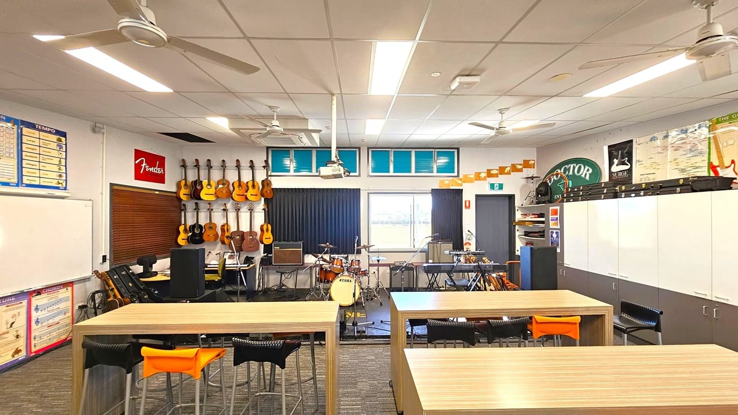 A music classroom with several guitars hanging on the left wall, beneath a "Fender" sign. In the centre, a drum kit is set up with microphones and amplifiers nearby. Keyboards and other musical equipment are arranged around the room. The right side features white cabinets with posters above. Tables and stools are in the foreground. The ceiling has fluorescent lights and ceiling fans, and a window at the back lets in natural light.