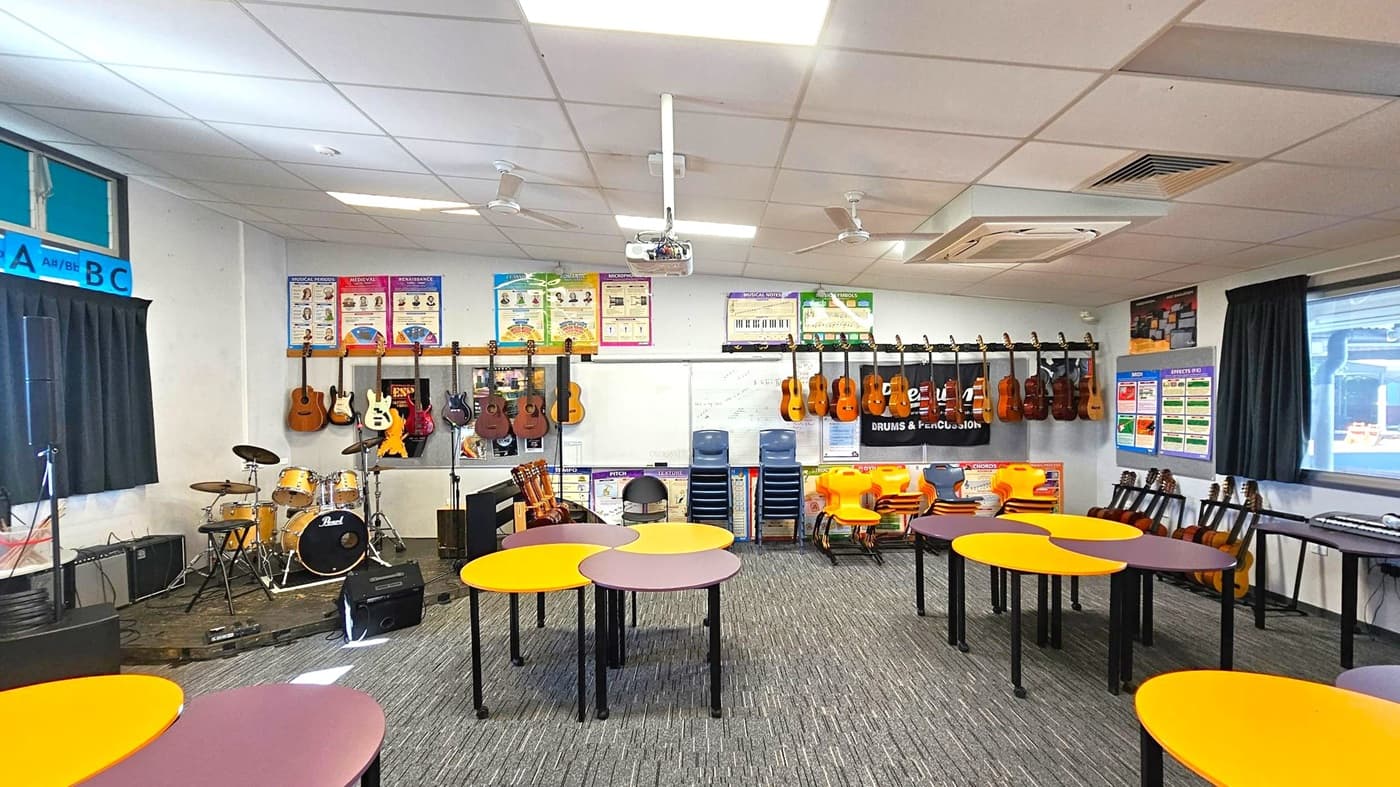 A music classroom with a variety of instruments and educational materials. On the left, a drum set and several guitars are displayed. The back wall features posters and more guitars hanging. In the centre, there are round tables with orange and purple tops. On the right, stacked chairs and additional guitars are visible. The floor is carpeted, and the ceiling has fluorescent lights and fans. Large windows with black curtains are on the left and right walls.