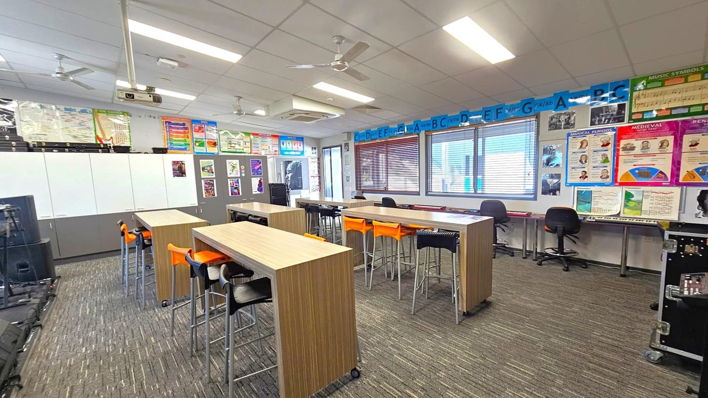 A classroom with a carpeted floor and a ceiling with fluorescent lights and ceiling fans. In the centre, there are long wooden tables with orange and black high stools. The left wall has white and grey cabinets with posters above them. The right wall features windows with blinds and more educational posters. At the back, there are musical instruments and equipment, including a keyboard and a speaker.