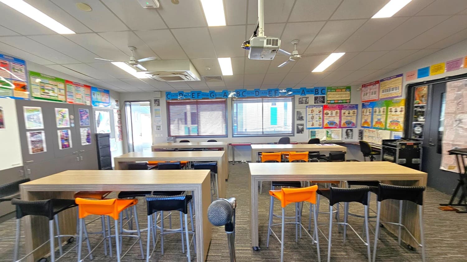 A classroom with a carpeted floor and a ceiling with fluorescent lights and a projector. In the foreground, there are tables with high chairs, some with orange backs. A microphone stand is visible. The walls are adorned with educational posters and a series of letters above the windows. Cabinets with more posters are on the left, and a door is visible in the background. The room has a bright and organised appearance.