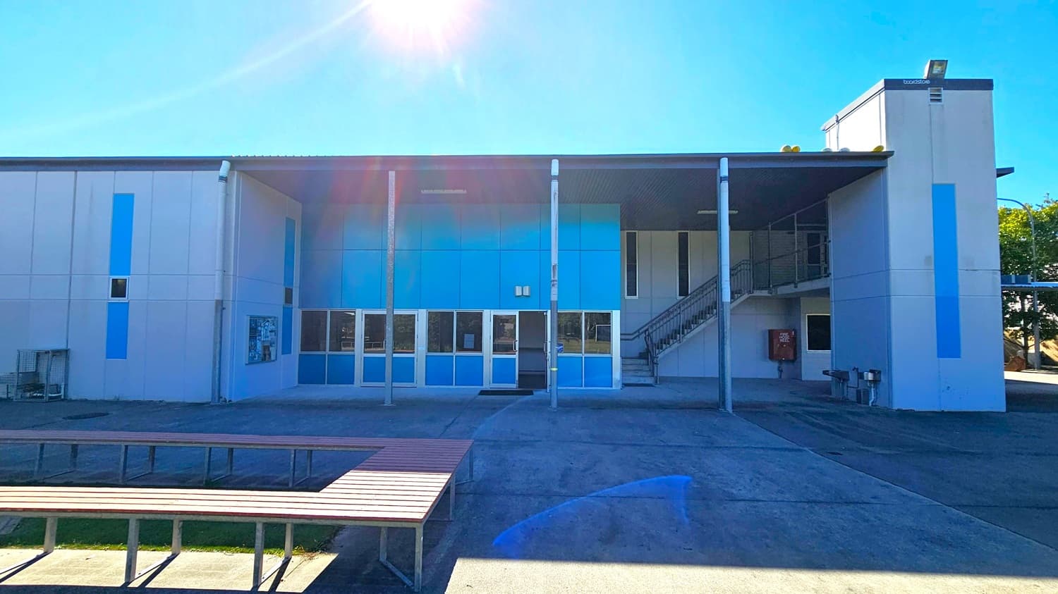 A two-storey building with a flat roof and a modern design. The facade features large blue panels and multiple windows. A staircase is visible on the right side, leading to the upper level. In the foreground, there are wooden benches arranged in a U-shape on a concrete surface. The sun is shining brightly, casting shadows on the ground. The building has a clean, minimalist appearance with a few small trees visible on the far right.