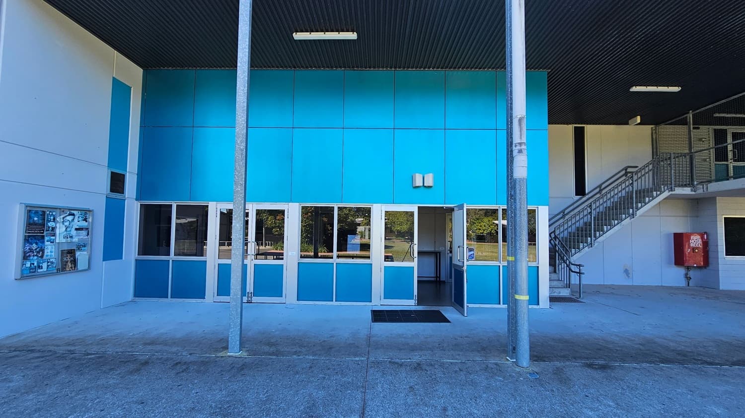A building exterior with a blue and white facade. In the centre, there are glass doors, one of which is open, leading inside. A metal pole stands in front of the entrance. On the left, a notice board displays various posters. On the right, a staircase with metal railings ascends to an upper level, and a red fire hose reel is mounted on the wall. The ceiling is dark with a light fixture, and the ground is concrete.
