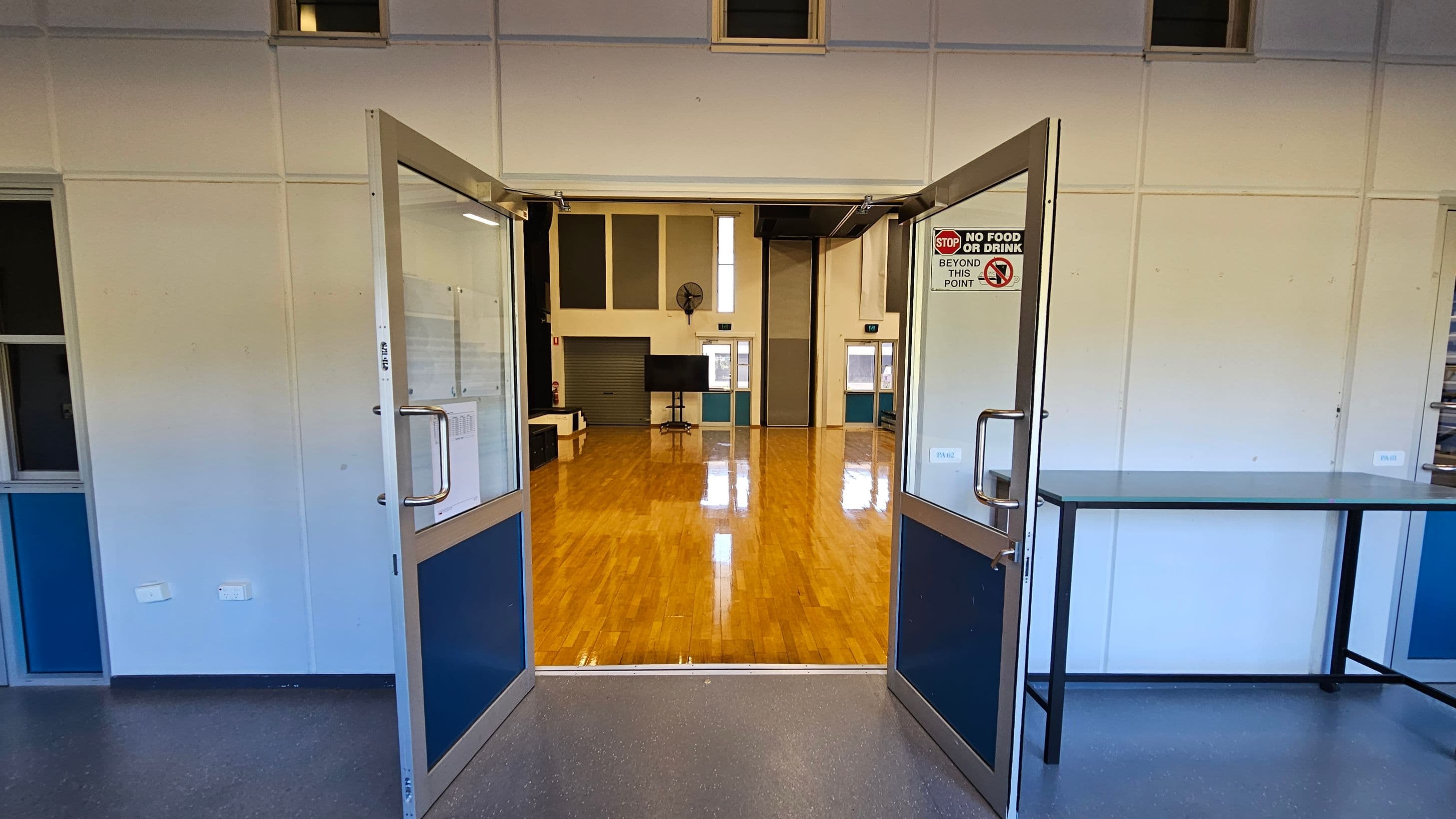 Double doors with glass panels are open, leading into a large room with a polished wooden floor. On the right, a sign on the door states "No food or drink beyond this point." Inside the room, there are two small windows and a fan on the far wall, along with a television on a stand. The walls are light-coloured with some darker panels. A table with a metal frame is positioned on the right side of the entrance. The ceiling is high with small windows near the top.