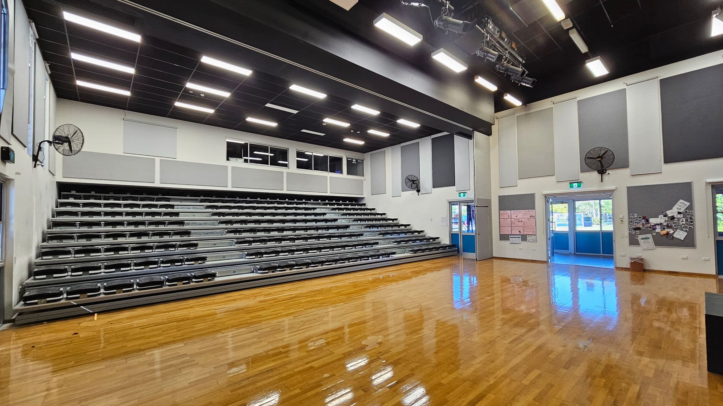 A spacious room with a polished wooden floor. On the left, there are tiered metal bleachers with several rows of seating. The walls are adorned with large grey and white acoustic panels, and there are wall-mounted fans. The ceiling is black with rectangular lights and stage lighting fixtures. On the right, there are double doors with glass panels leading outside, and a notice board with various papers pinned to it.