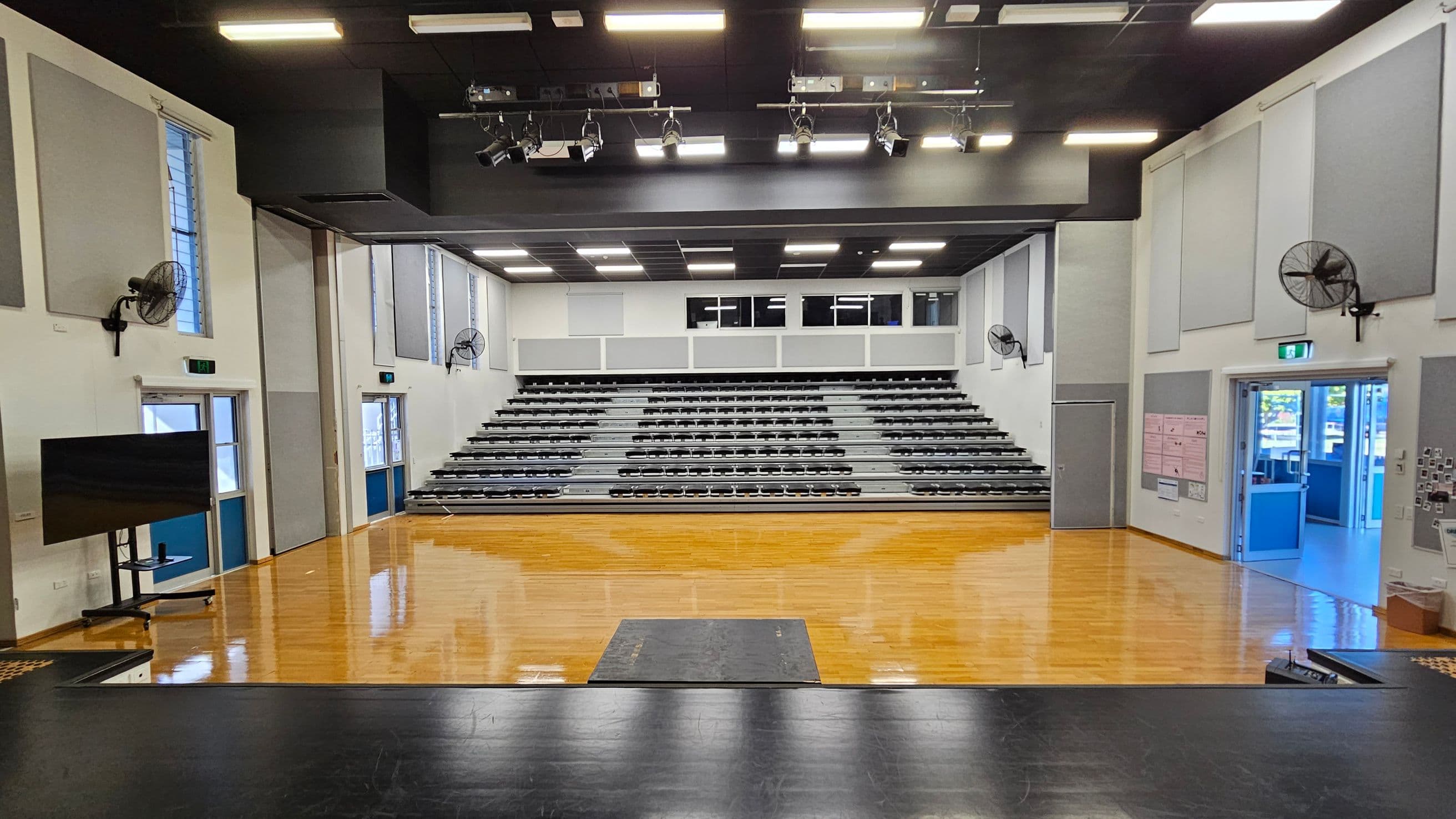 A spacious auditorium with a polished wooden floor. In the centre, there are tiered grey seating rows. The ceiling is equipped with multiple lights and stage equipment. On the left, a large screen is mounted on a stand near a set of double doors. On the right, another set of double doors is open, leading to a brightly lit corridor. Wall-mounted fans are visible on both sides, and the walls are lined with acoustic panels.